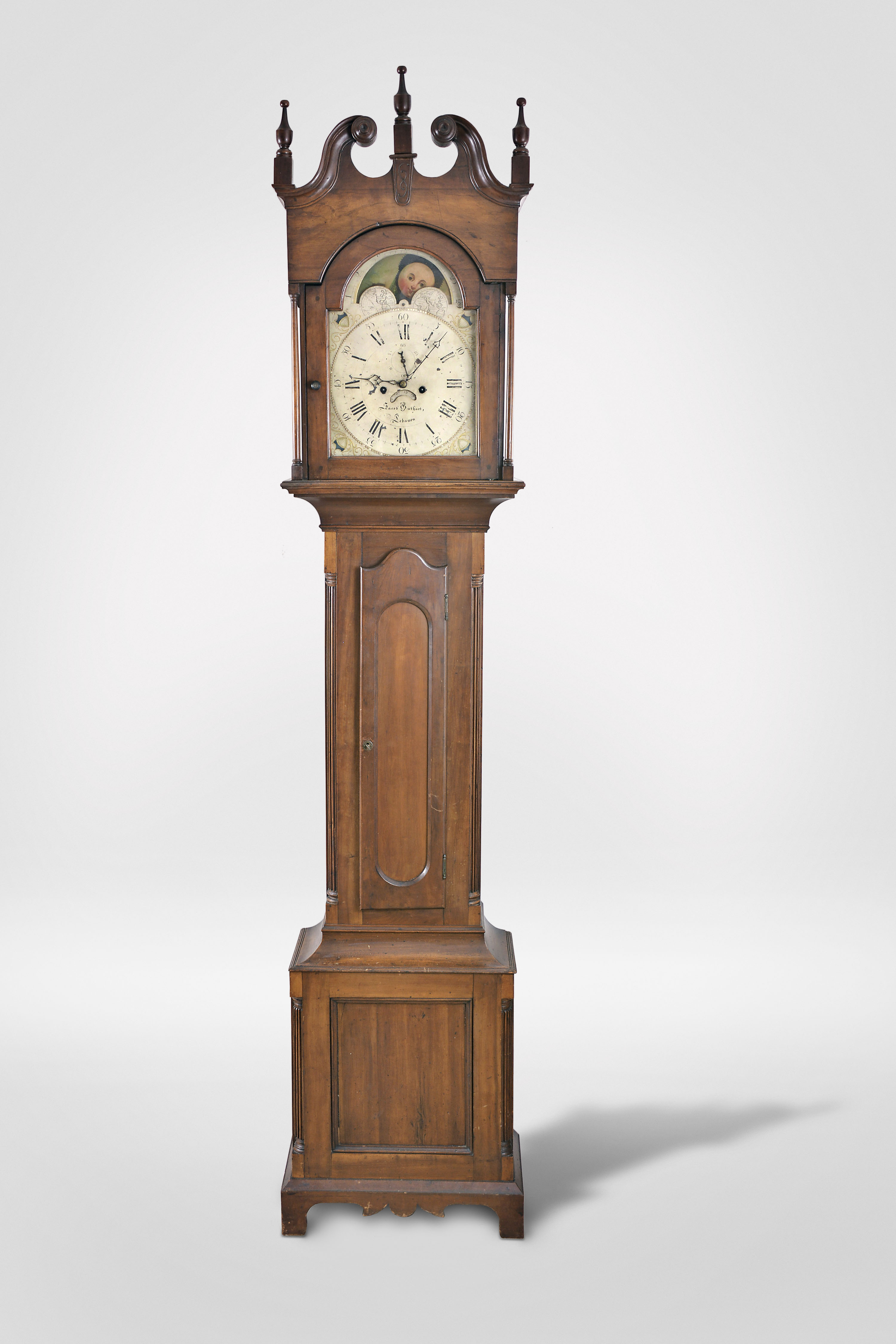 Jacob Guthart, Lebanon, Penn., tall clock, 8 day, time and strike, weight driven movement in a handsome cherry case with broken arch pediment set with concentric turned rosettes, three wooden finials, full columns flanking hood, shaped trunk door flanked by fluted quarter columns over base with matching quarter columns and recessed panel on bracket feet with cutout skirt. The painted metal is signed by maker and has moon phase, calendar and seconds bit. The dial was made and signed by "NOLEN'S MANUFACTY BOSTON ". — Antique Clocks Price Guide