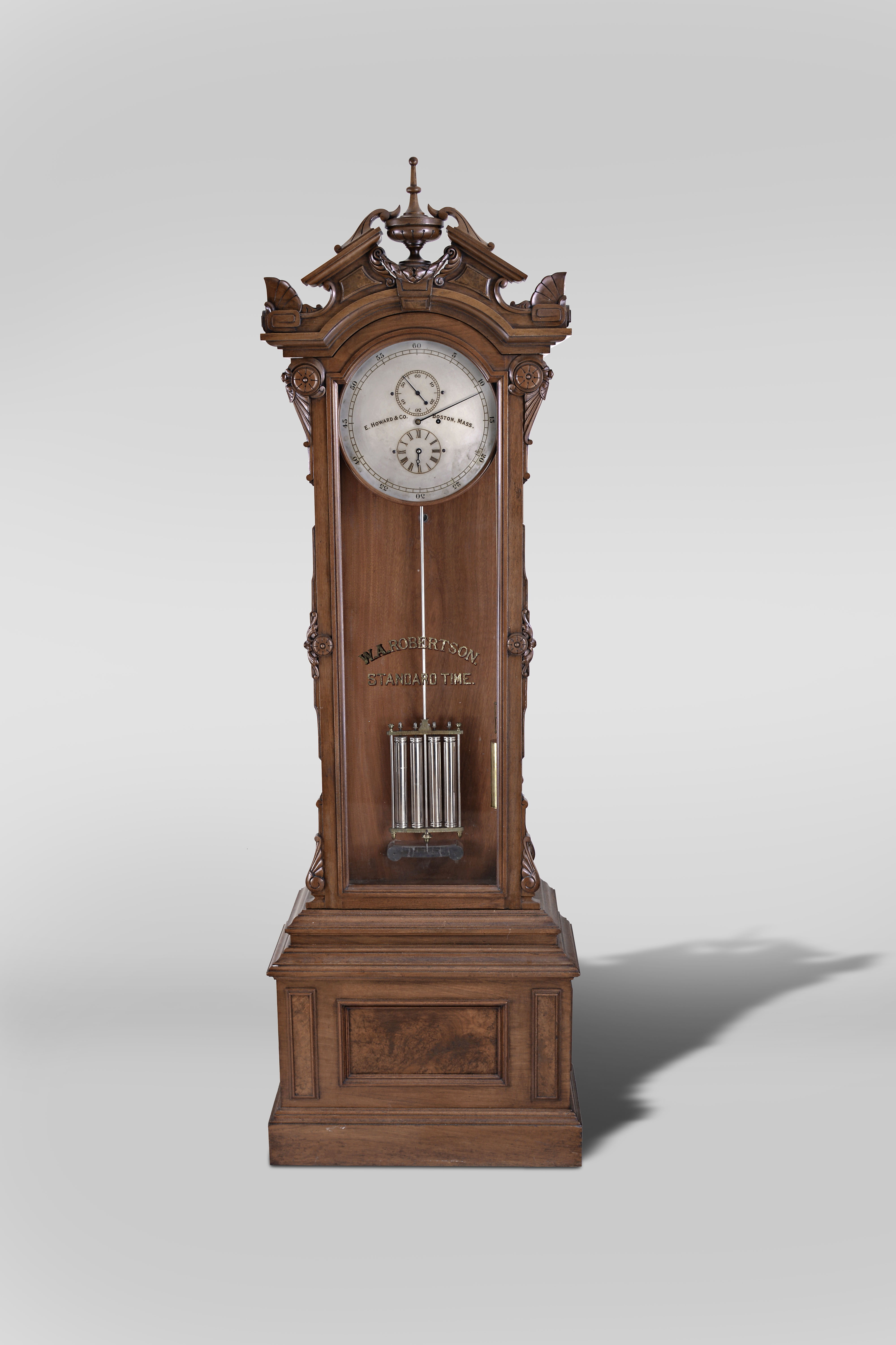 E. Howard & Co., Boston, "No. 61" astronomical regulator, the renaissance revival style case with molded stepped base and burl veneered panels to the front and sides, and with cyma and cavetto molded top, supporting the upper case with glazed door and sides, the door with applied, stylized Grecian foliate carvings, and simple eight lobe roundels, arched cornice molding surmounted by a broken pitched pediment with central urn and laurel festoon, and the corners with stylized palmette form acroterion angularium above stylized, flattened volutes, door glass with reverse painted and gilded "W. A. Robertson Standard Time", silvered astrnomical dial signed "E. Howard & Co. Boston, Mass.", blued steel hands, damascened brass week going movement with jewelled pallets, mercury pendulum with four nickel plated steel jars, brass frame, and nickel plated rod, silvered beat scale, brass clad weight, and heavy cast iron bracket attached to the case back, supporting both the pendulum and the movement — Antique Clocks Price Guide