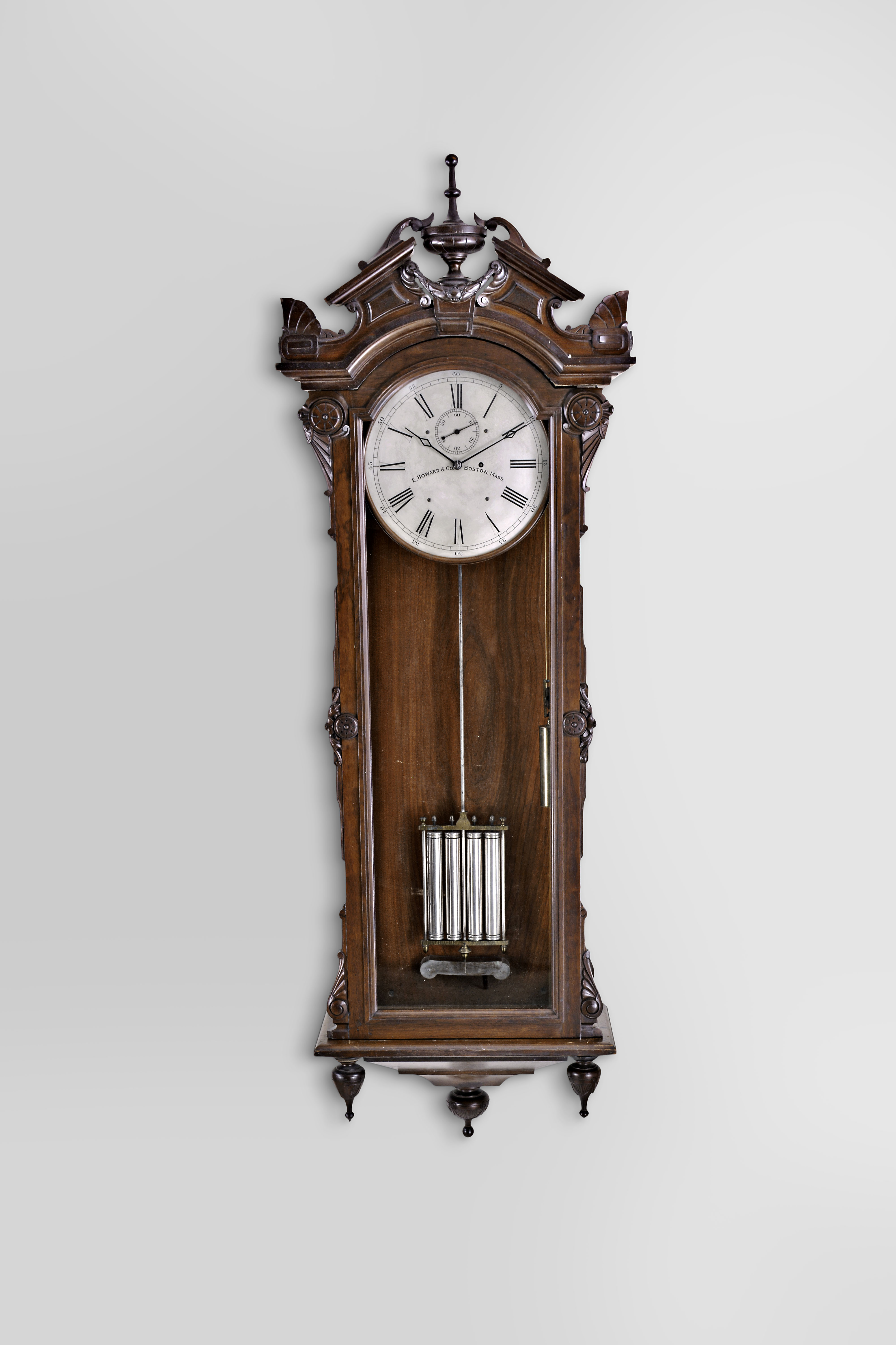 E. Howard & Co., Boston, "No. 60" hanging regulator, the renaissance revival style case with glazed door and sides, the door with applied, stylized Grecian foliate carvings, and simple eight lobe roundels, base with two turned, carved drops flanking a tapered, stepped bracket terminating in a large, shallow cavetto molding with turned and carved drop, the top with arched cornice molding surmounted by a broken pitched pediment with central urn and laurel festoon, and the corners with stylized palmette form acroterion angularium above stylized, flattened volutes, silvered dial signed "E. Howard & Co. Boston, Mass.", blued steel hands, damascened brass week going movement with jewelled pallets, mercury pendulum with four nickel plated steel jars, brass frame, and nickel plated rod, silvered beat scale, brass clad weight, and heavy cast iron bracket attached to the case back, supporting both the pendulum and the movement — Antique Clocks Price Guide
