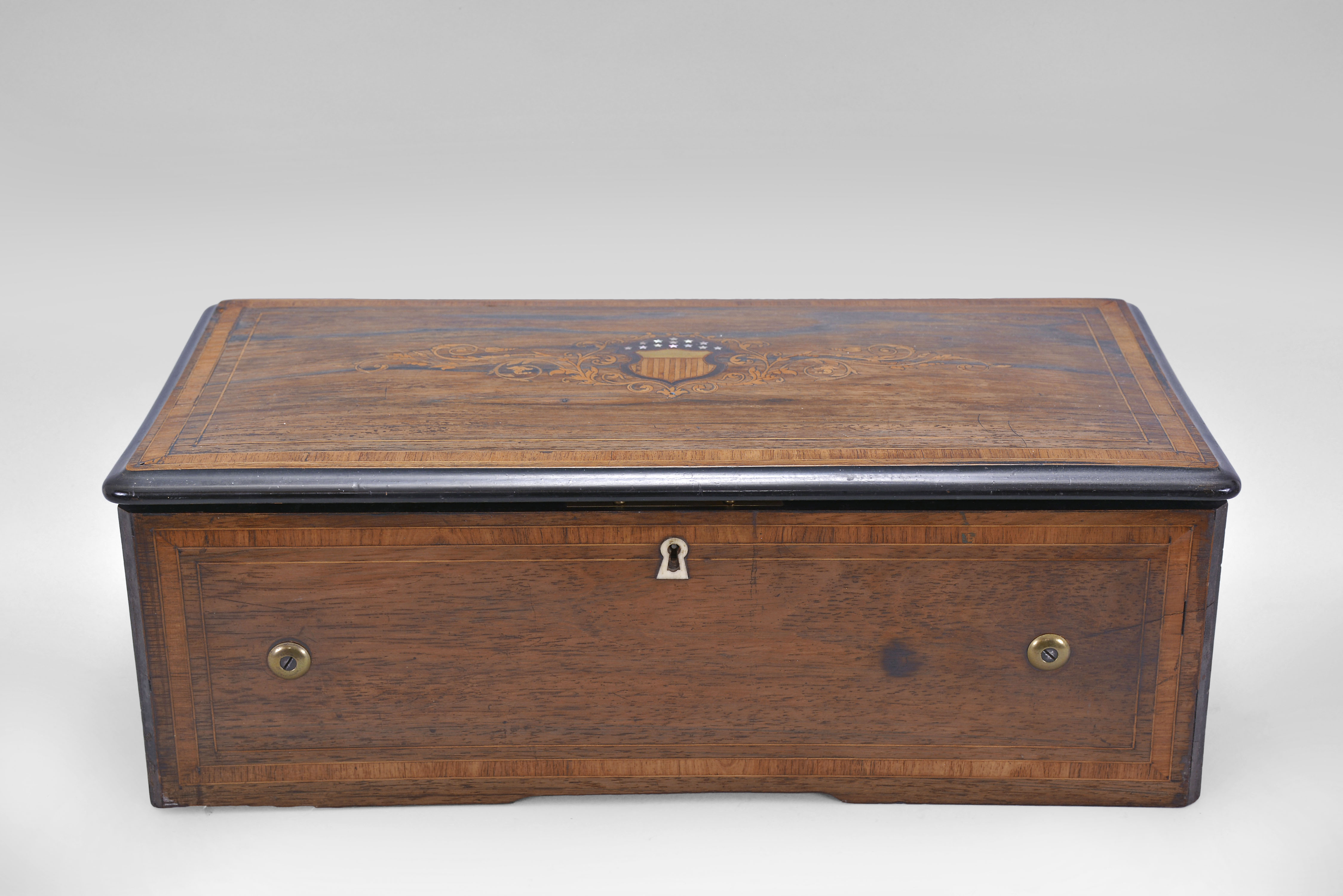 Switzerland, music box, playing 8 tunes with a 12 inch cylinder playing on a two-piece comb with 92 teeth and six tuned bells, mounted in a box with rosewood veneered top with marquetry scrolls, a striped shield and 13 Mother of Pearl stars, the front with string inlay, original tune card mounted under the lid listing the patriotic themed songs. — Antique Clocks Price Guide