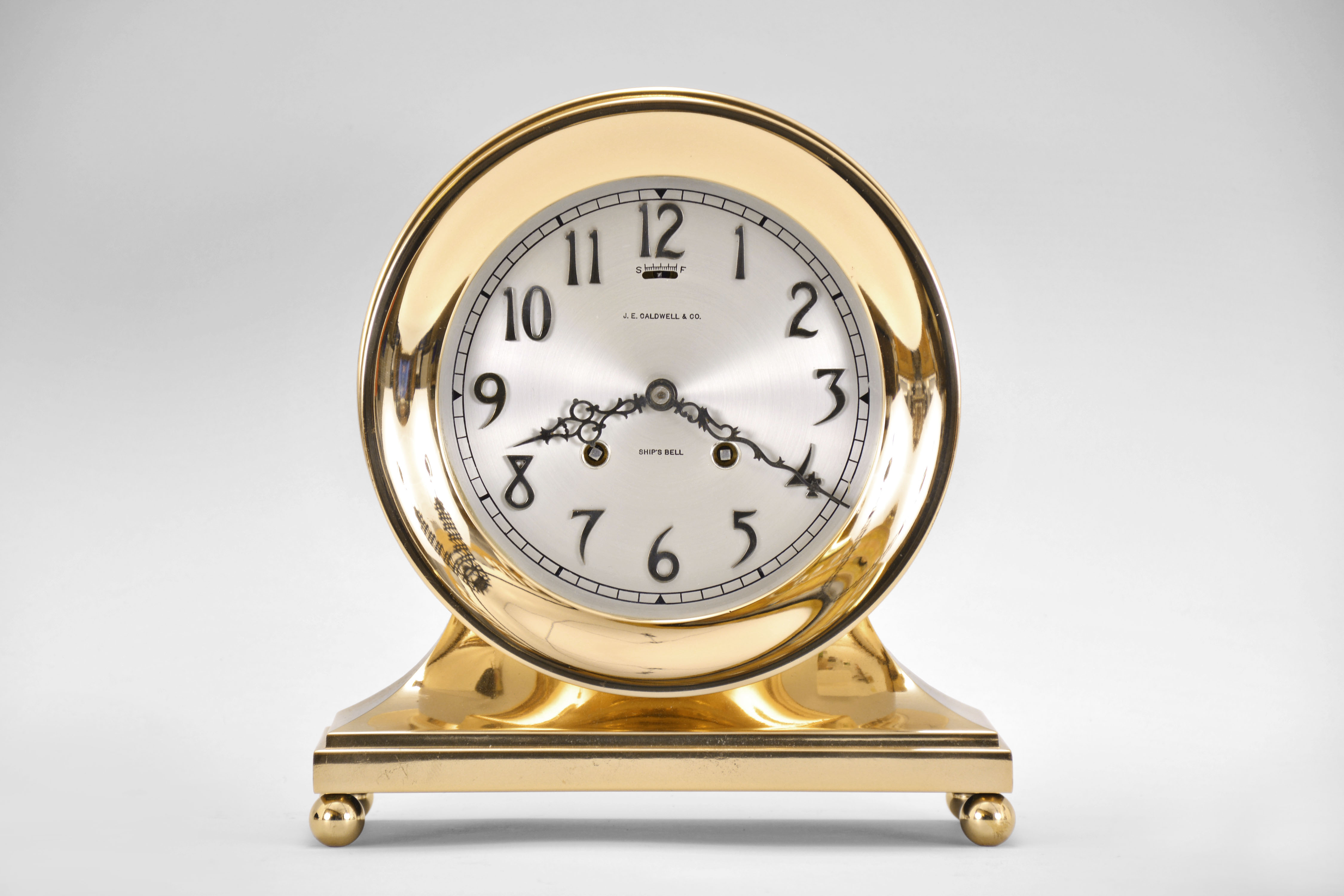 Chelsea Clock Co., Boston, Mass., "Constitution" shelf clock for J.E. Caldwell with six inch dial, polished brass case on an incurved base with ball feet, signed Arabic numeral silvered special dial with applied numerals also marked "Ships Bell", blued steel hands, 8 day, time and strike movement with lever escapement, serial #131632 matching on bottom and base and movement including Chelsea marked key — Antique Clocks Price Guide