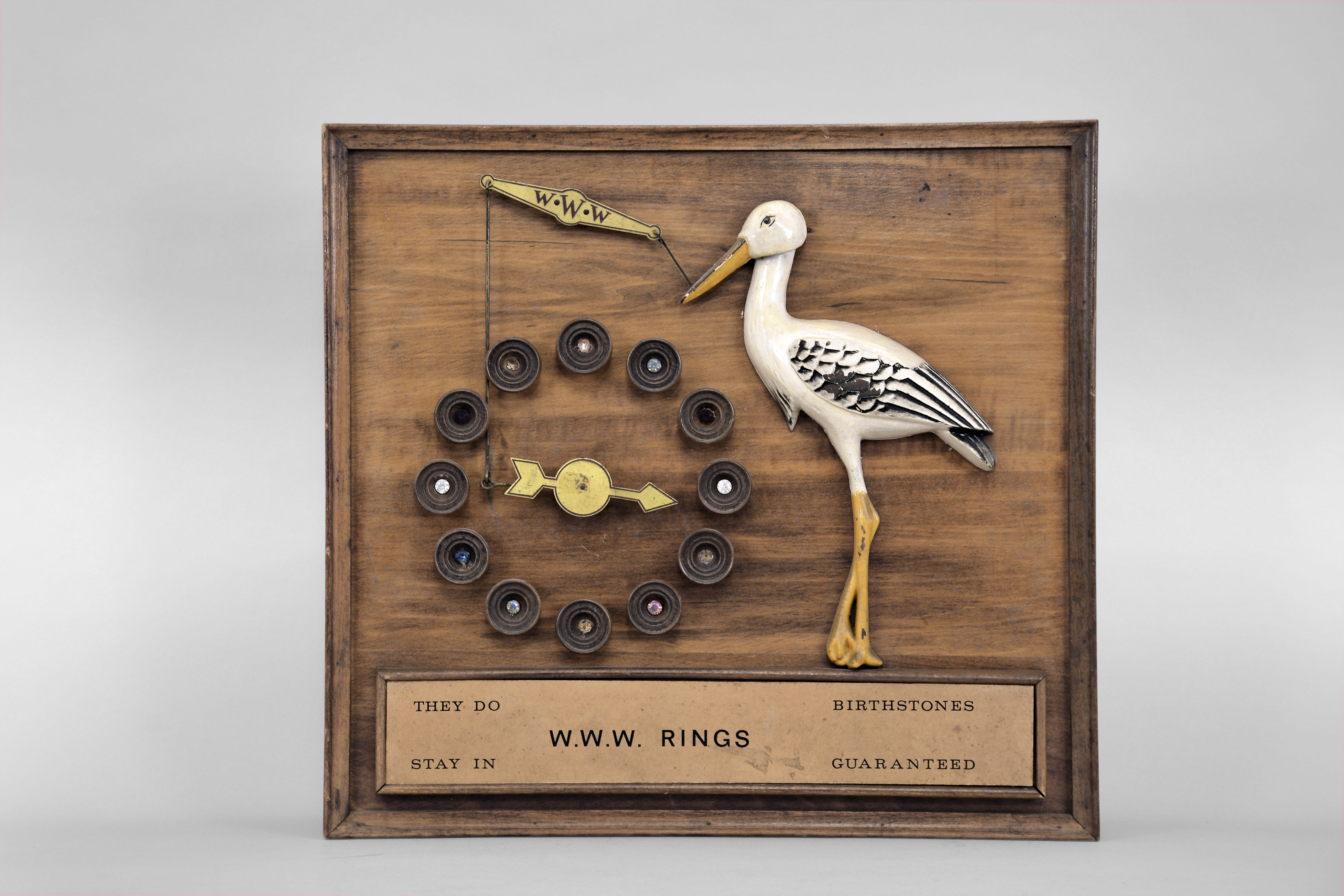 The Attracto Company, Philadelphia, Penn., jeweler's advertising display, spring driven pendulum mechanism in a wood case driving a carved nodding stork figure and rotating arrow pointing to circular display of birthstones, the plaque on the front reading "They Do Stay In / Birthstones Guaranteed" and "W.W.W. Rings ". — Antique Clocks Price Guide