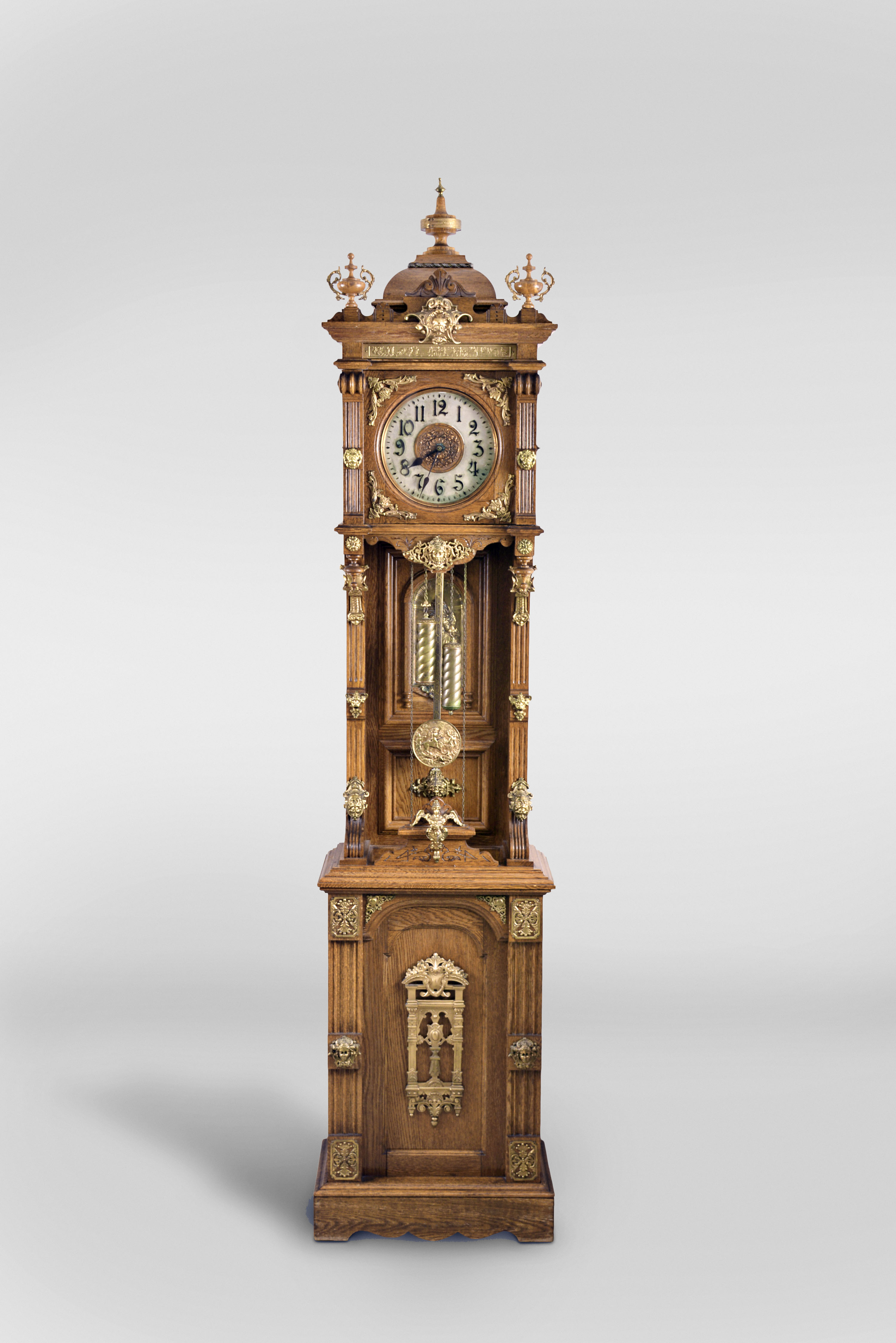 Ansonia Clock Co., New York, New York, "Antique Standing" tall clock, 8 day, time and strike, weight driven movement in an open well oak case with cast brass ornamentation and two brass rope turned weights and brass clad wood end pendulum rod with embossed pendulum bob featuring putti and lion scene. The 10" metal dial has silvered chapter ring, copper center and raised numerals. — Antique Clocks Price Guide