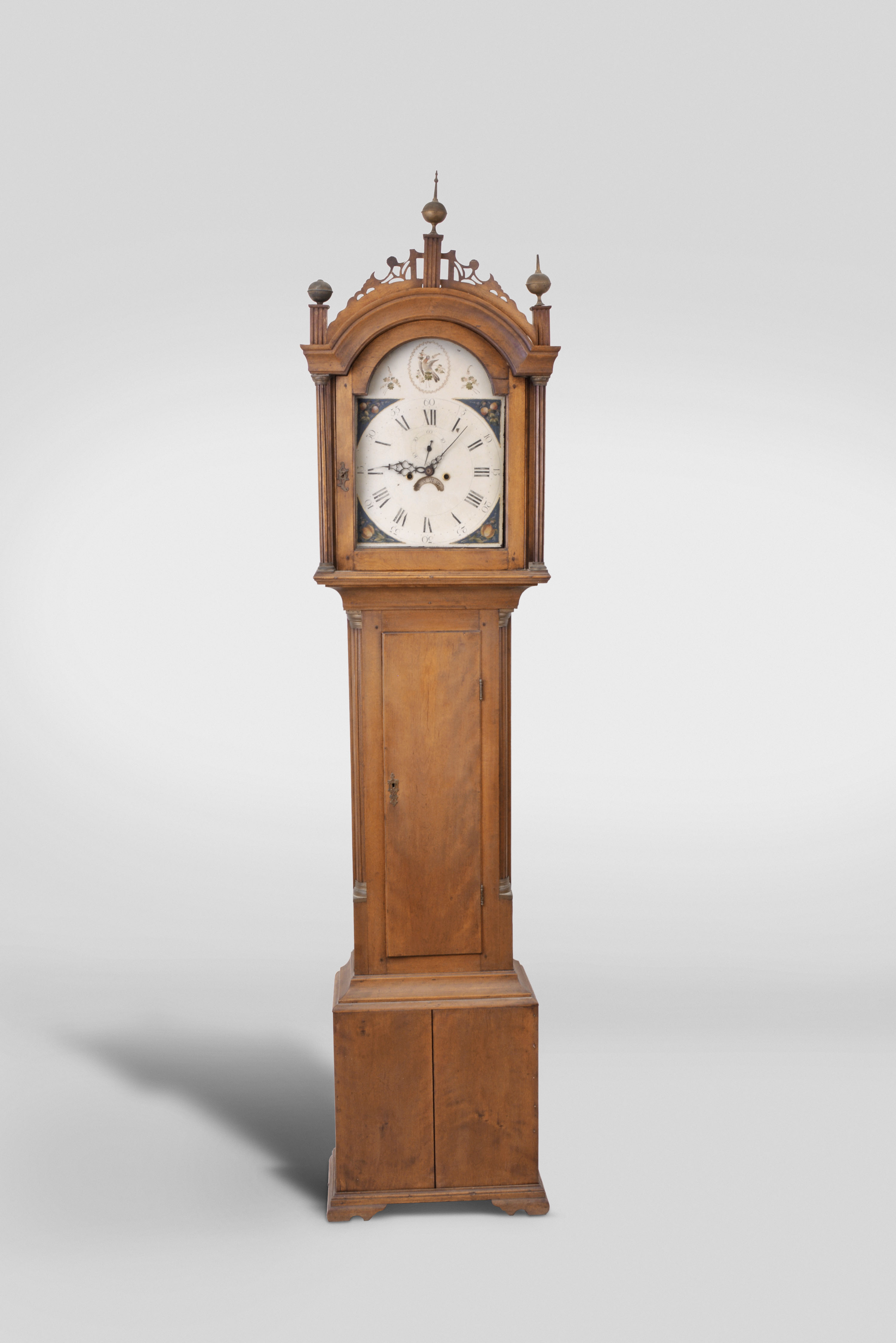 New England tall clock, 8 day, time and strike, weight driven movement in a cherry case with fretwork top, three brass finials and fluted columns on hood over trunk with fluted corner columns with brass capitals over base resting on ogee feet — Antique Clocks Price Guide
