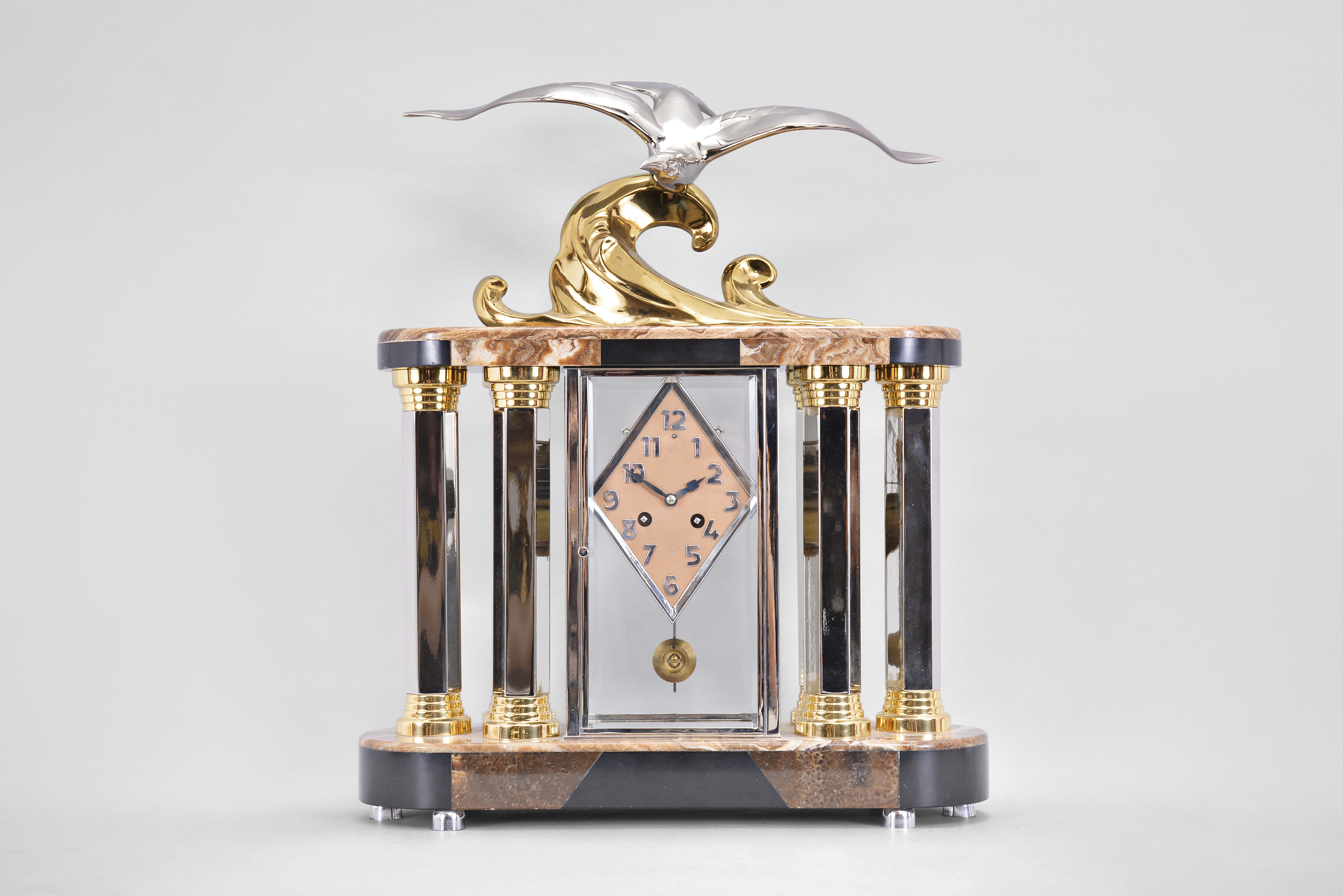 Michel Decoux, a marble and polished metal mantel clock with gull ornament, the oblong base and top with brown, variegated marble veneers over contrasting black slate base and panels, joined portico style by six hexagonal, polished chrome columns with polished, gilt capitals and bases, and flanking a polished chrome crystal regulator case, surmounted by a polished, gilt wave form casting and polished chrome gull, signed "Decoux", diamond shaped Arabic numeral metal dial with blued steel hands, and 8 day Pendule de Paris movement, striking on a bell — Antique Clocks Price Guide