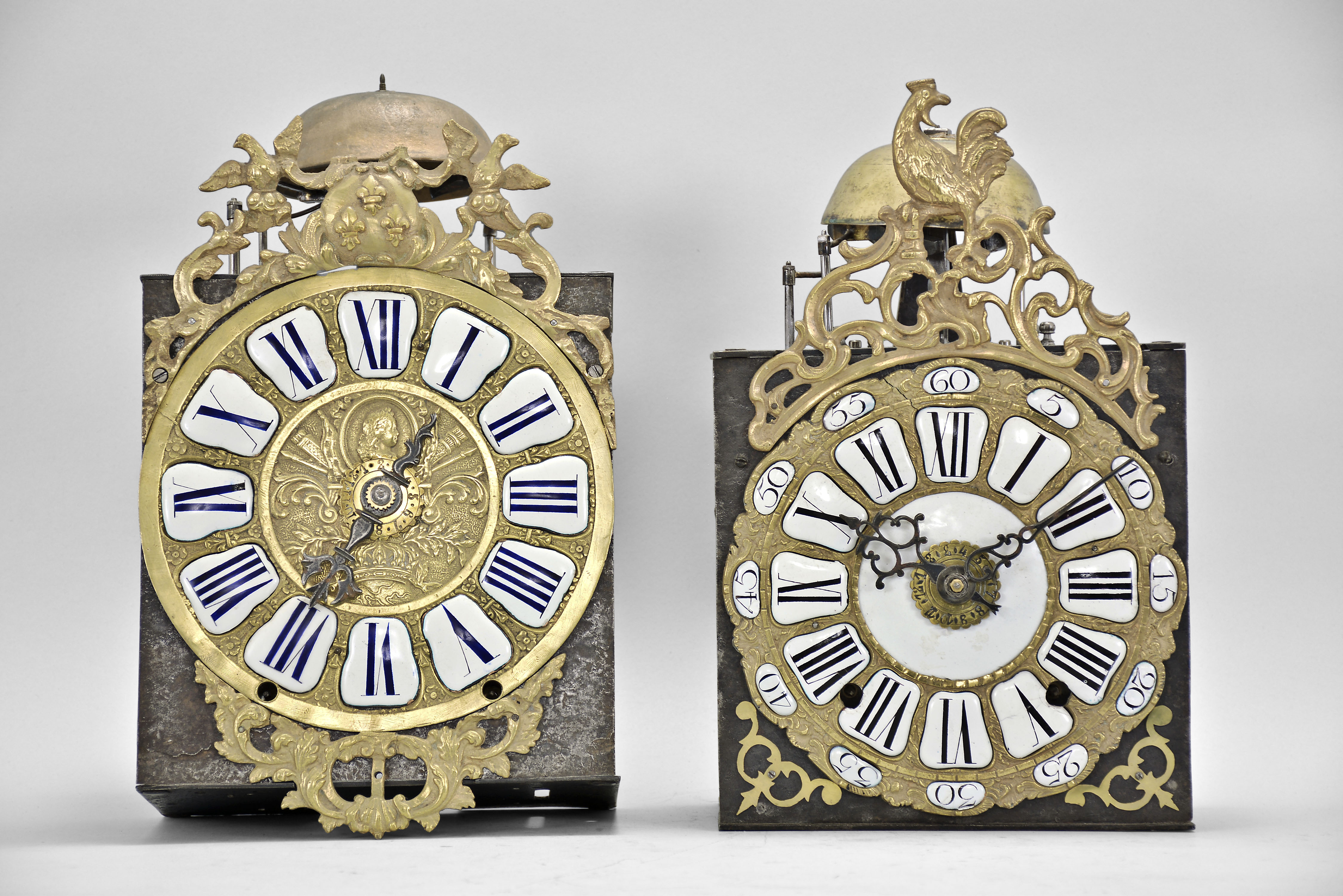 Clocks- 2 (Two) Comtoise with Cartouche Numerals: (1) France, bronze-front Comtoise clock, 8 day, time and strike with alarm, weight driven movement with verge escapement, thread suspension with rear-hanging pendulum in an iron post and plate frame, cast brass dial with royal symbols including a crown and profile of Louis XV, white enamel cartouche numerals with blue lettering, topped by cast fronton featuring fleur de lis and the backward facing Gallic rooster, applied lower casting, and single iron hand, mid 18th Century. (2) France, bronze-front Comtoise clock, 8 day, time and strike with alarm, weight driven movement with verge escapement, thread suspension with rear-hanging pendulum in a posted frame, 25 white enamel panels inset within cast brass dial, two iron hands, pieced brass spandrels, and topped by cast fronton with backward facing Gallic rooster, mid 18th Century. — Antique Clocks Price Guide