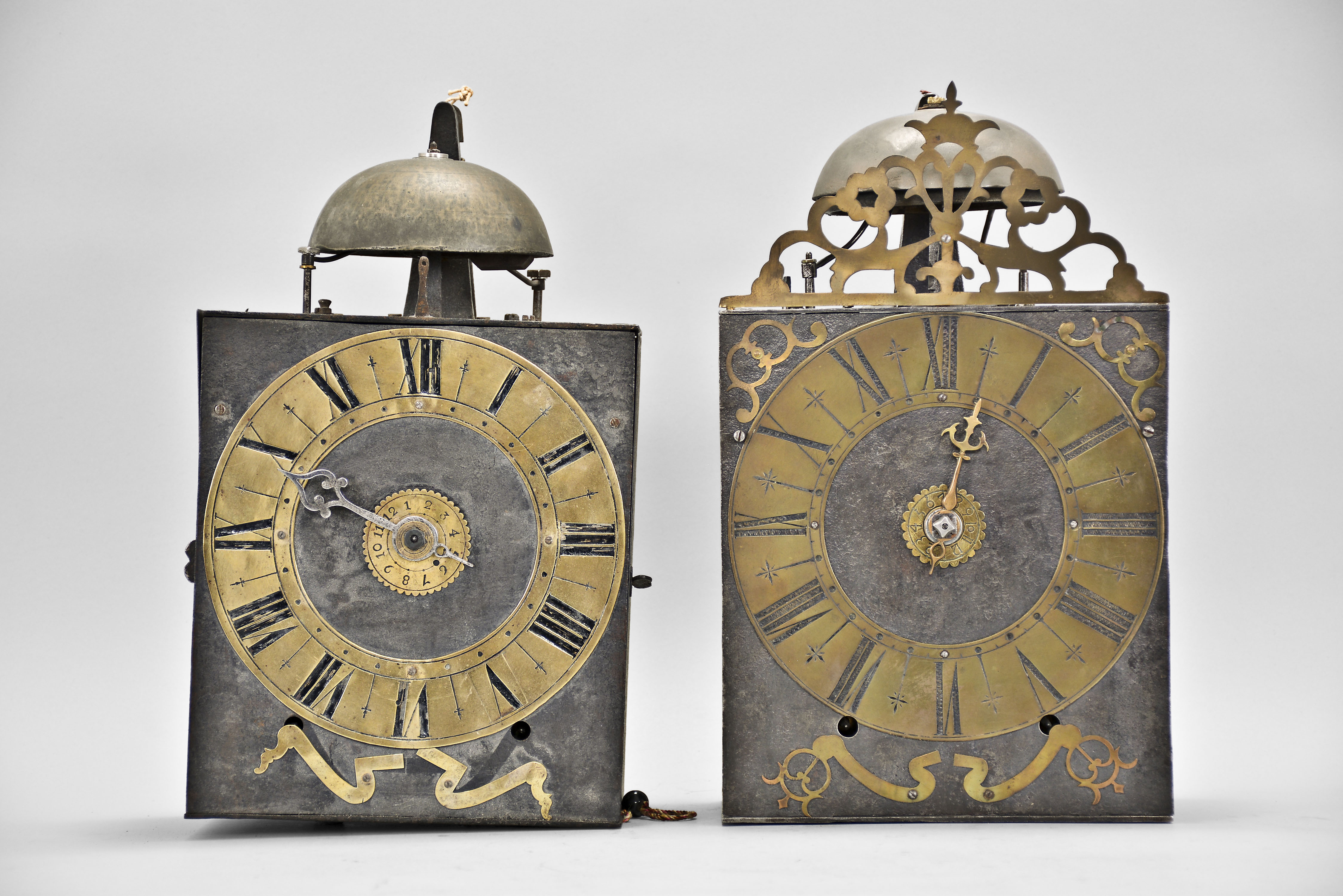 Clocks- 2 (Two) Early Comtoise: (1) France, Comtoise clock, 8 day, time and strike, weight driven movement with verge escapement, thread suspension with rear-hanging pendulum in a iron post and plate frame, brass chapter ring with engraved Roman numerals, pivoted winding arbor shutters, and single iron hand, first half of the 18th Century. (2) France, Comtoise clock, 8 day, time and strike with alarm sounding on a second bell, weight driven movement with verge escapement, thread suspension with rear-hanging pendulum in a iron post and plate frame, brass chapter ring with engraved Roman numerals, applied brass spandrels and pivoted winding arbor shutters, and single brass hand, first half of the 18th Century. — Antique Clocks Price Guide