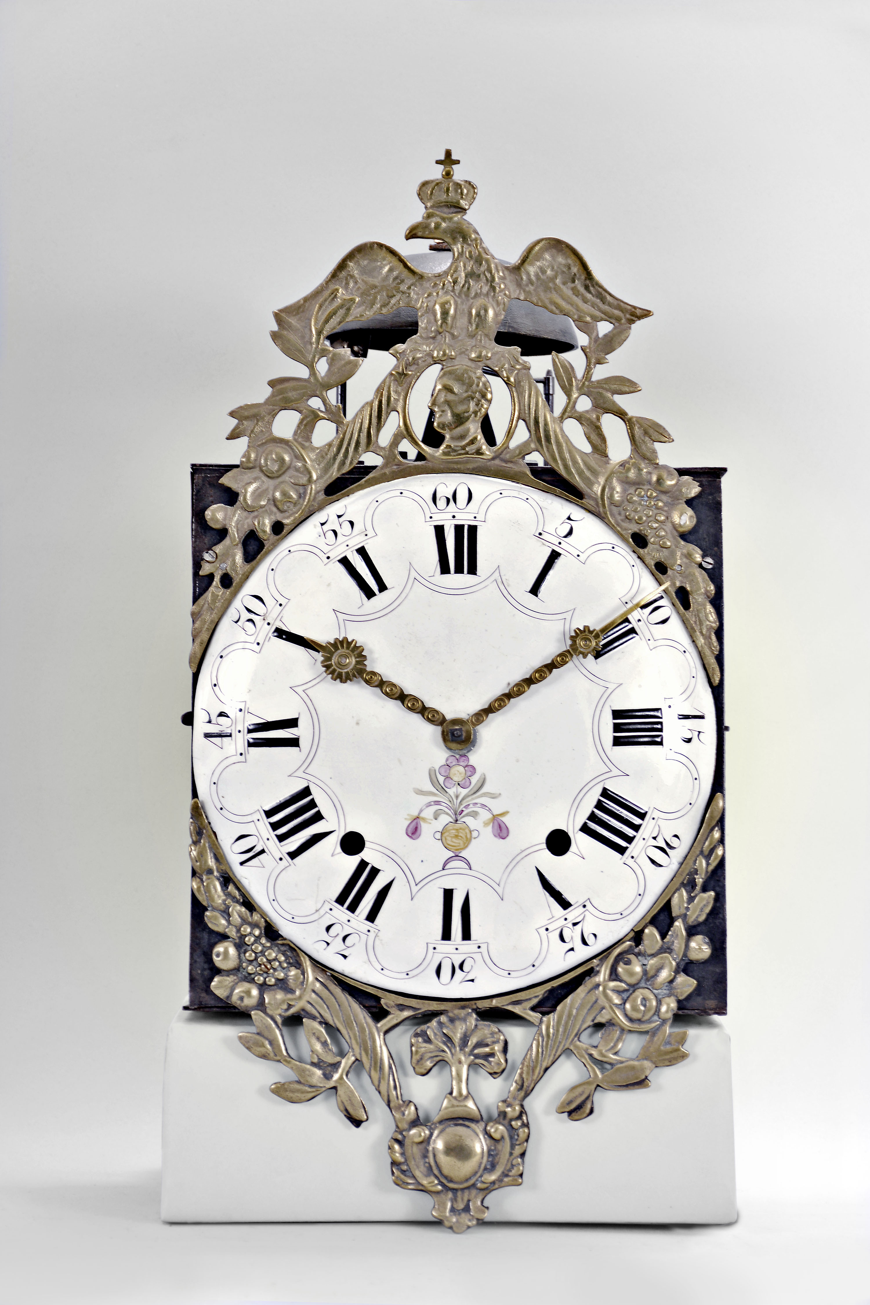 France, Comtoise wall clock, 8 day, time and strike, weight driven movement with verge escapement, thread suspension with front-hanging pendulum in an iron post and plate case with white enamel convex Roman numeral arcaded dial with Arabic minutes, polychrome floral decoration above the 6 o'clock position, two brass sunburst hands, cast brass fronton, the lower portion with a central tree flanked by cornucopias, the top with a profile of Napoleon below the crowned eagle symbol of the Empire. — Antique Clocks Price Guide