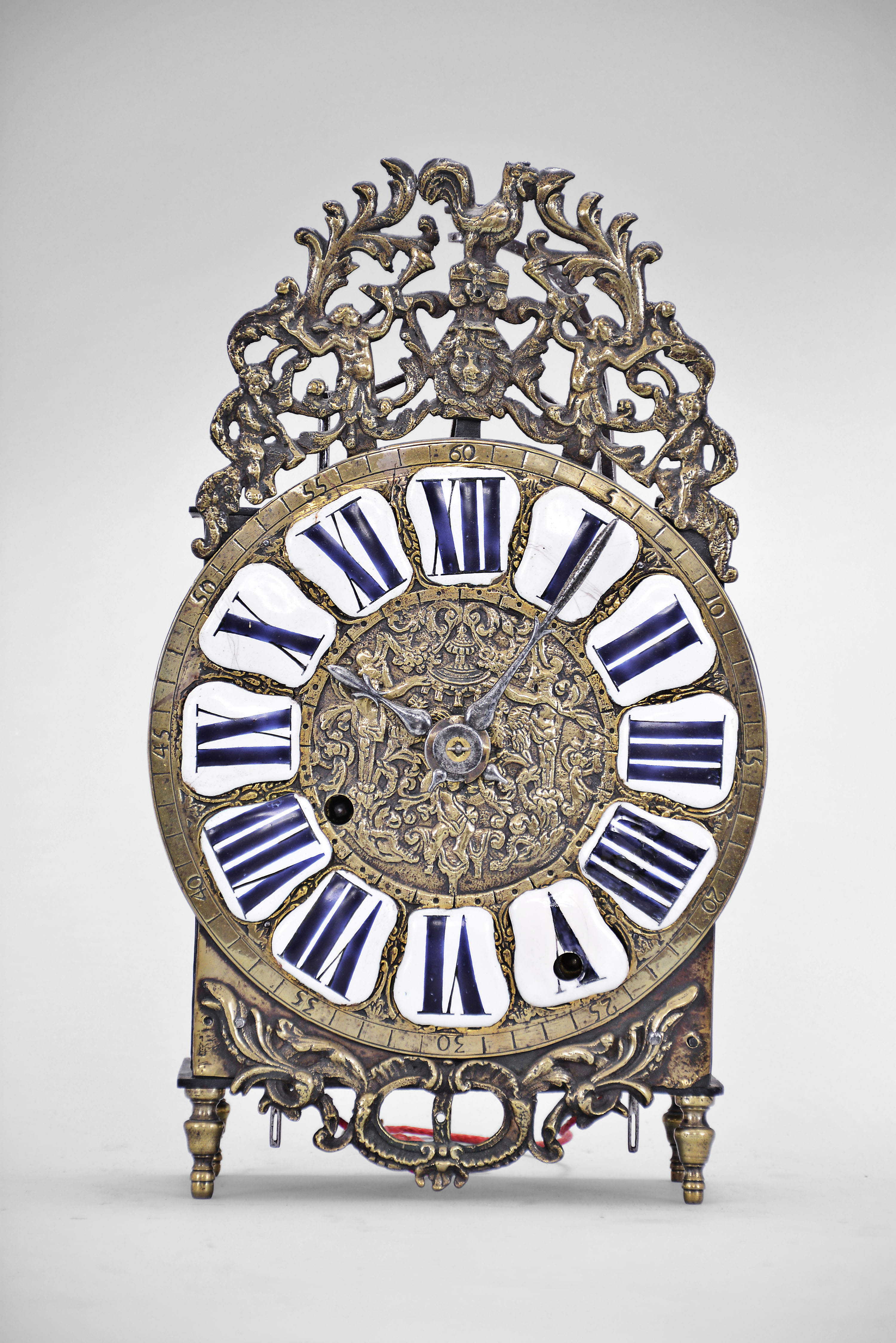 France, Comtoise lantern clock, 8 day, time and strike, weight driven condensed movement with verge escapement, wire suspension with rear-hanging pendulum in an iron posted case with hammered brass doors, cast brass dial with inset white enamel cartouches with blue Roman numerals, brass outer ring with Arabic numerals every 5 minutes, two steel hands, topped by cast bronze fronton featuring trumpeters and a forward facing Gallic rooster, all sitting on four turned brass feet. — Antique Clocks Price Guide