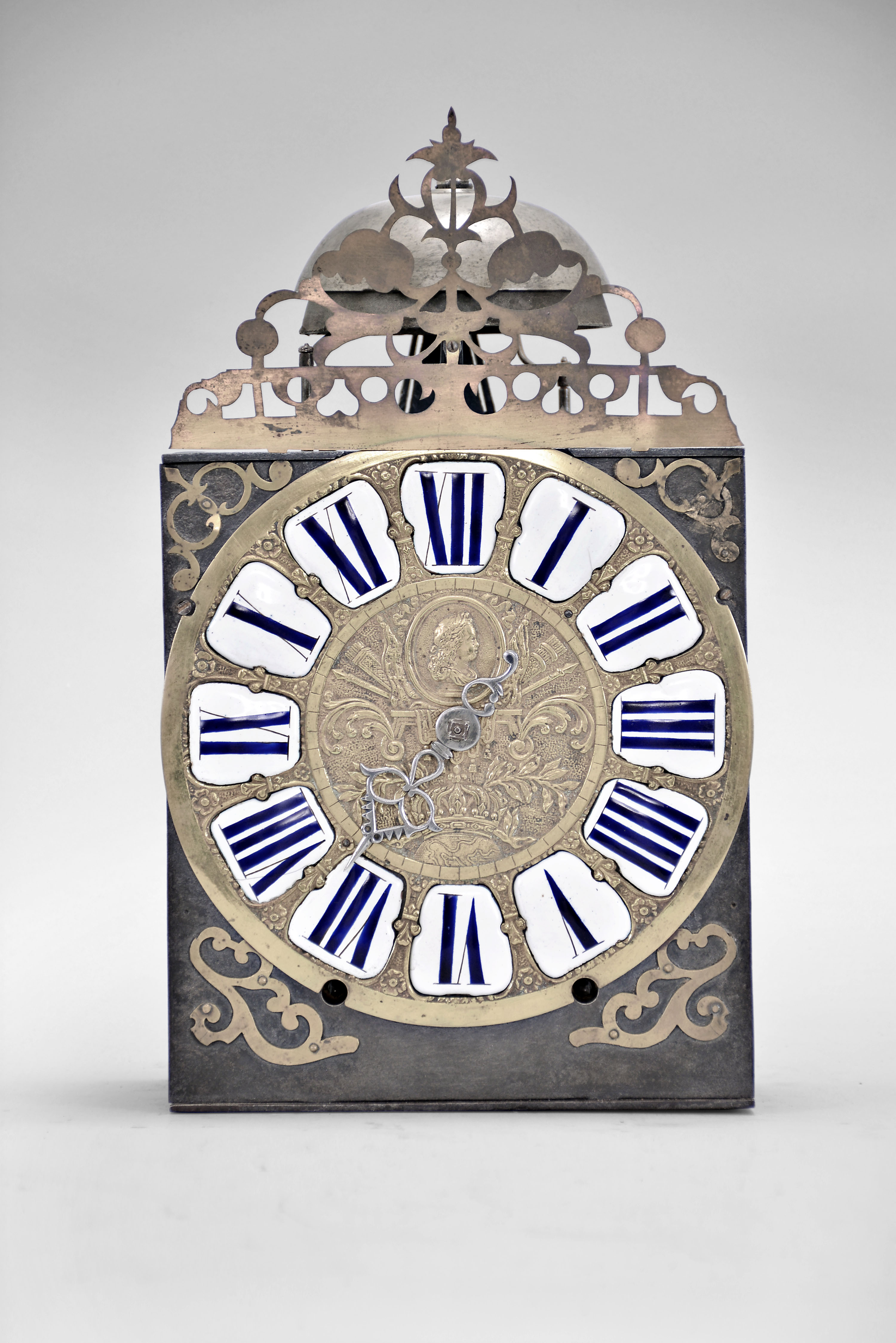 France, Comtoise clock, 8 day, time and strike, weight driven movement, verge escapement, thread suspension with rear-hanging pendulum in an iron post and plate frame, cast brass dial with royal symbols including a crown resting on the earth and profile of a young Louis XV or his son The Dauphin, white enamel cartouches with blue Roman numerals, single iron hand, four flat brass spandrels, topped by pierced flat brass fronton, mid 18th Century. — Antique Clocks Price Guide