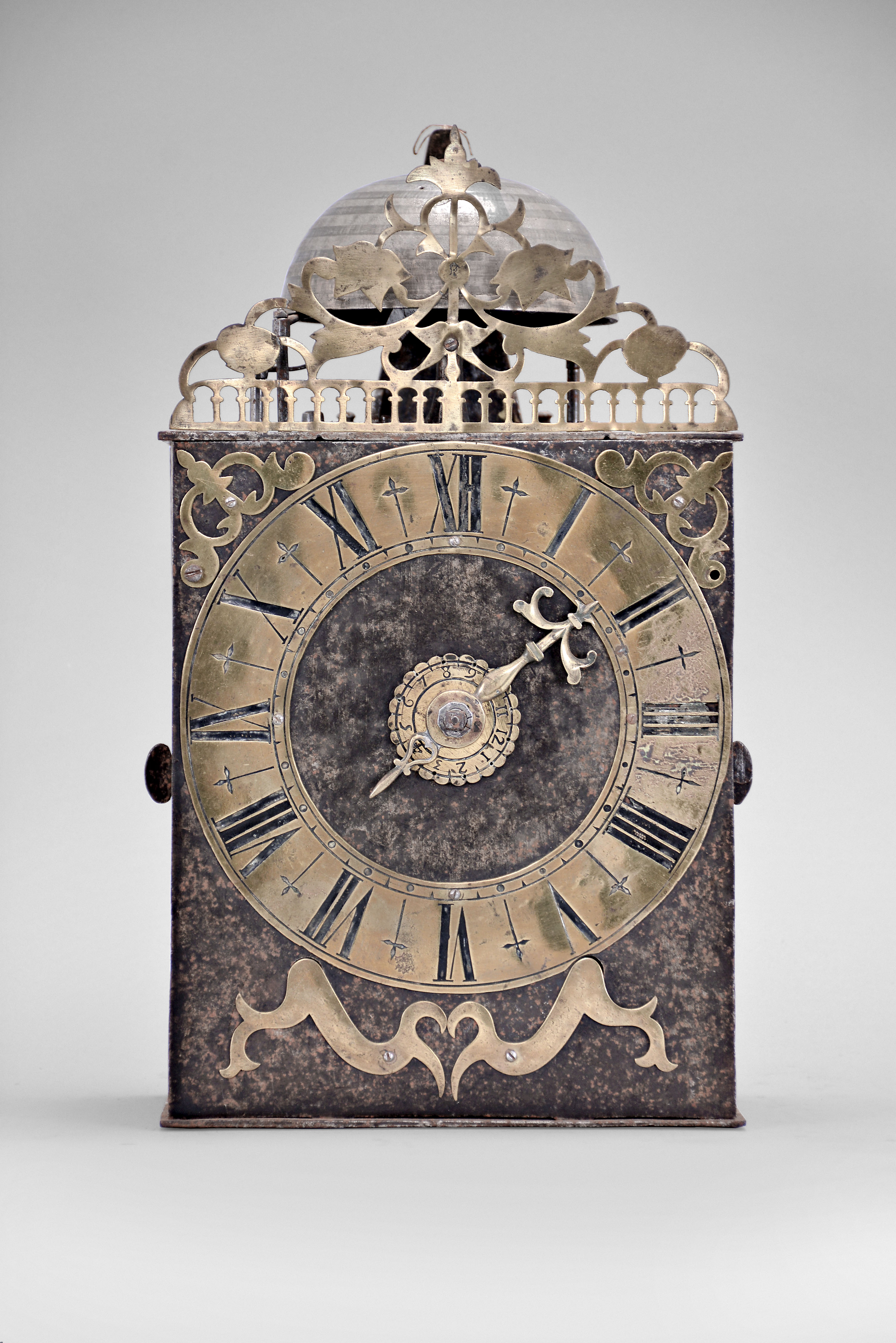 France, Comtoise clock, 8 day, time and strike with alarm, weight driven movement with verge escapement, thread suspension with rear-hanging pendulum in a iron post and plate frame, brass chapter ring with engraved Roman numerals, alarm dial with Arabic numerals, single brass hand, applied brass spandrels and pivoted winding arbor shutters, topped by flat pierced brass fronton with trademark. — Antique Clocks Price Guide