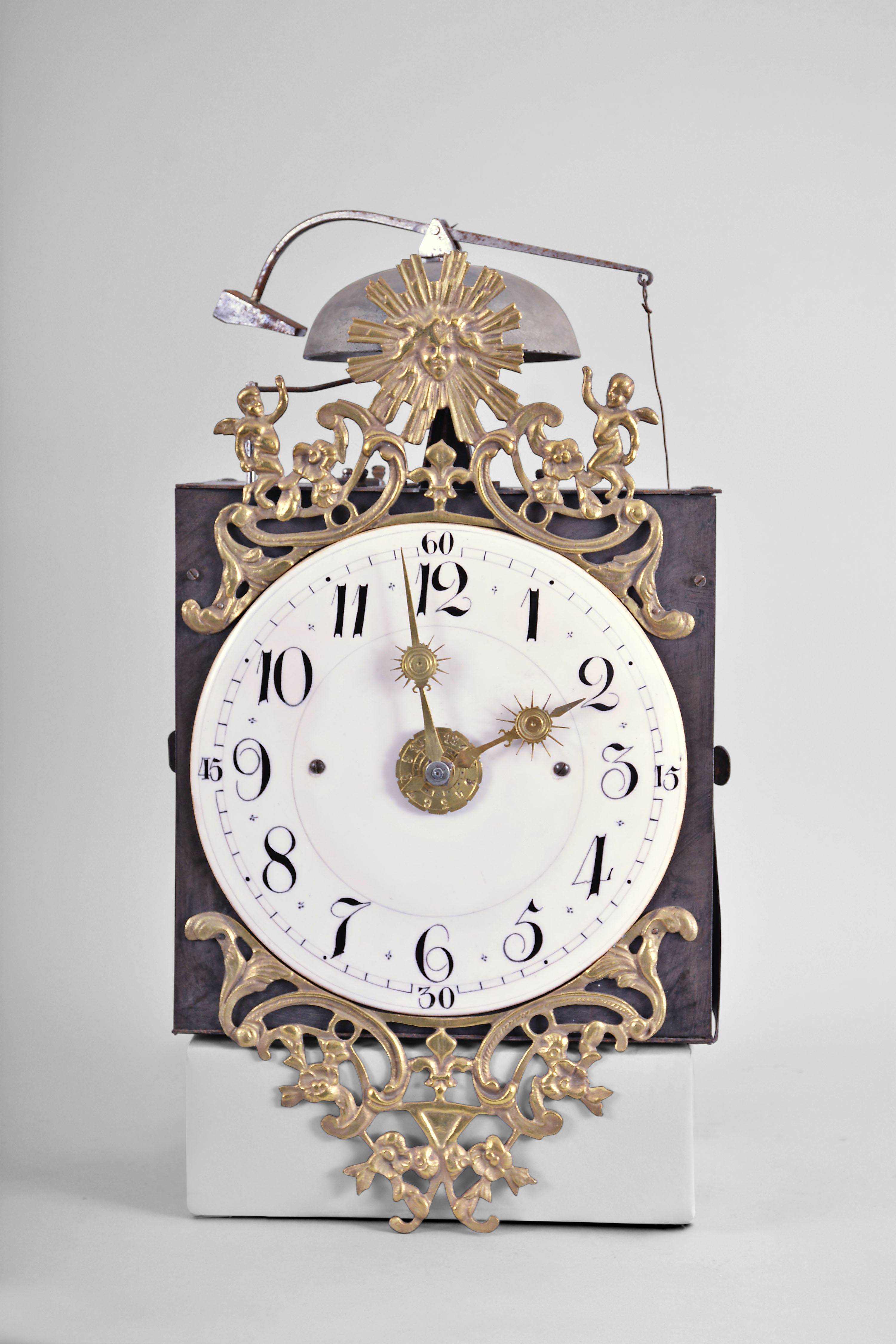 France, Restoration Period bronze-front Comtoise clock, 8 day, time and strike with alarm, weight driven movement with rear winding arbors, verge escapement with thread suspension and front-hanging pendulum in an iron posted frame, convex faience dial with Arabic numerals for the hours and smaller numerals at 15 minute intervals, alarm ring with Arabic numerals, two brass hands with sun bursts, cast brass fronton with central sun image above a single fleur de lis and flanked by putti, lower fronton casting with flowers and fleur de lis. — Antique Clocks Price Guide