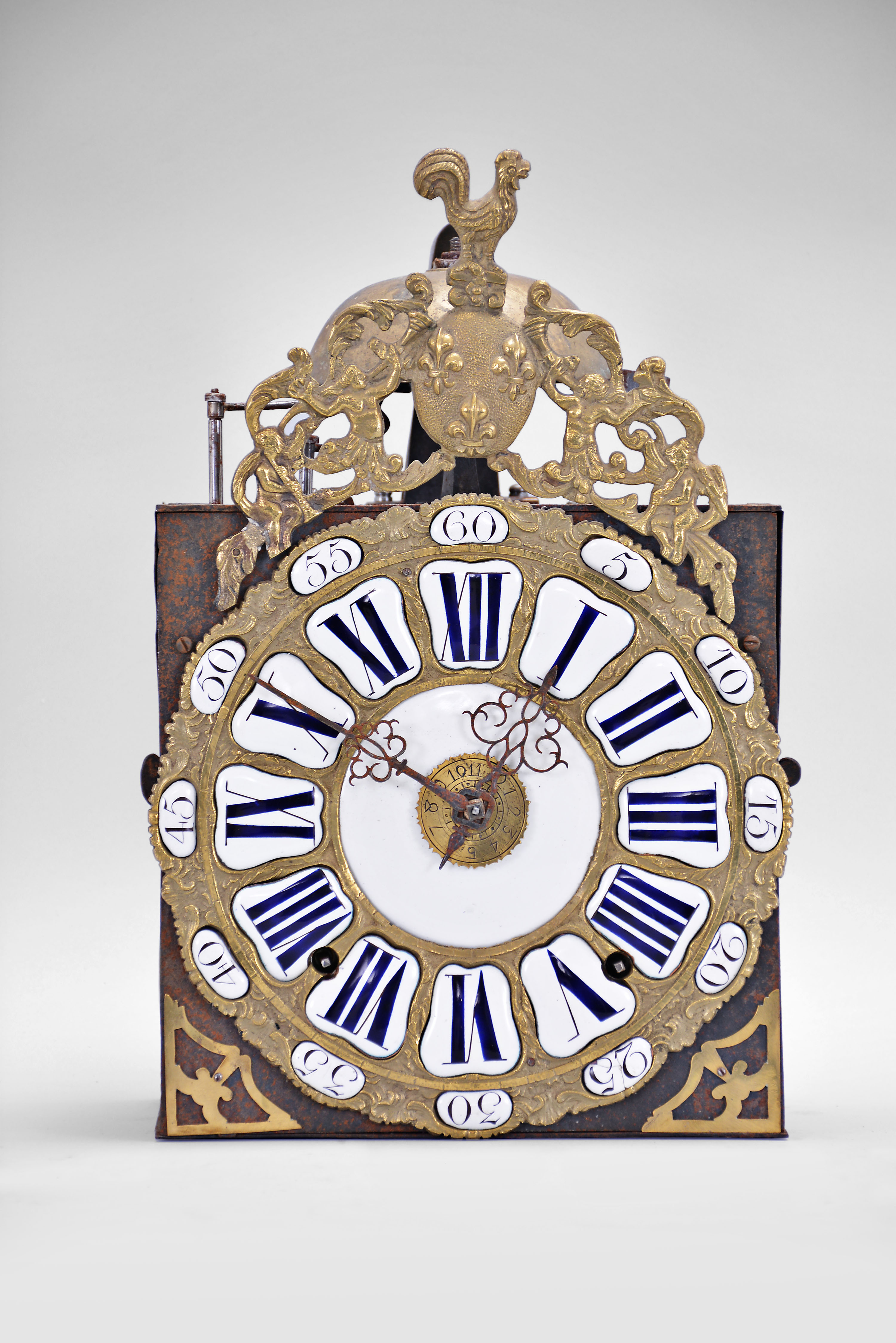 France, bronze-front Comtoise clock, 8 day, time and strike with alarm, weight driven movement, verge escapement with thread suspension and rear-hanging pendulum in an iron post and plate frame, cast brass dial with inset white enamel cartouches with blue Roman numerals for the hours and black Arabic numerals for the minutes, brass alarm ring with Arabic numerals, two pierced steel hands, applied brass spandrels below the dial, and topped with a cast fronton featuring a forward facing Gallic rooster over a medallion with three fleur de lis. — Antique Clocks Price Guide