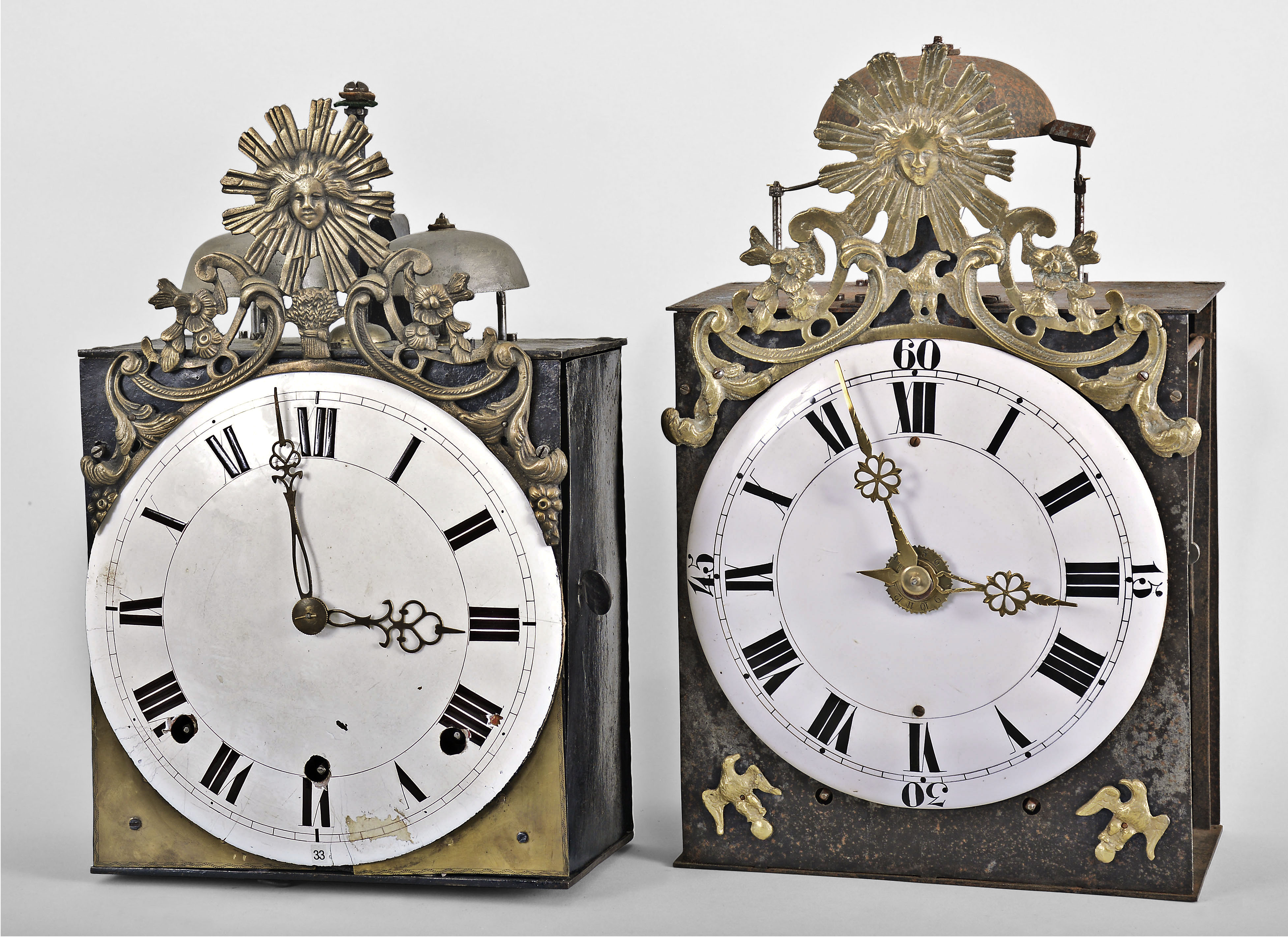 (1) France, Comtoise clock, 8 day, time and quarter strike, weight driven movement with verge escapement, thread suspension with rear-hanging pendulum in an iron post and plate frame, convex white enamel dial with Roman numerals for the hours, two pierced brass hands, cast bronze fronton featuring the sun and/or Apollo, flowers, and a sheaf of wheat, c1800; (2) France, month-running Comtoise clock, 30 day, time and strike with alarm, weight driven movement with verge escapement, thread suspension with front-hanging pendulum in a posted iron case, convex faience dial with Roman numerals for the hours, and Arabic numerals at the quarters, brass alarm ring with Arabic numerals, two brass hands, cast eagle spandrels, and cast fronton with central sun and eagle images, c1810. — Antique Clocks Price Guide