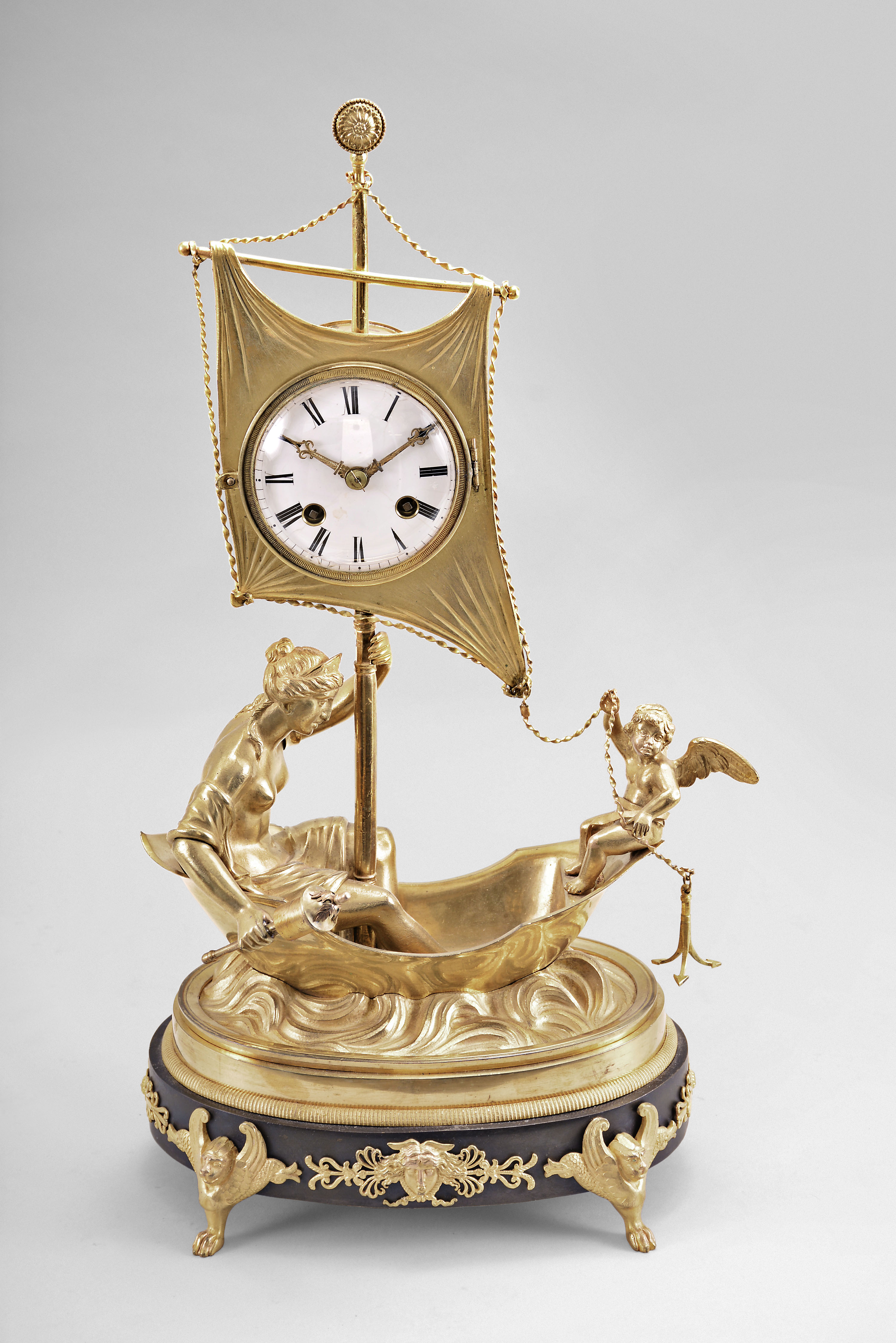 France, an unusual early 19th century gilt and patinated bronze figural clock, the oval base with four winged lion masks above paw feet, anthemion and palmette mounts, with central Medusa mask, and featuring two figures in a small sailboat, the rear with female figure, her left hand on the mast, and right hand holding what may be a torch, the bow with figure of cupid, his right hand raised, his left holding an anchor, the roman numeral white enamel dialmounted to tyhe sail, fancy gilt hands, two train pendule de Paris movement with silk thread suspension, striking on a bell, with associated oval base and dome — Antique Clocks Price Guide