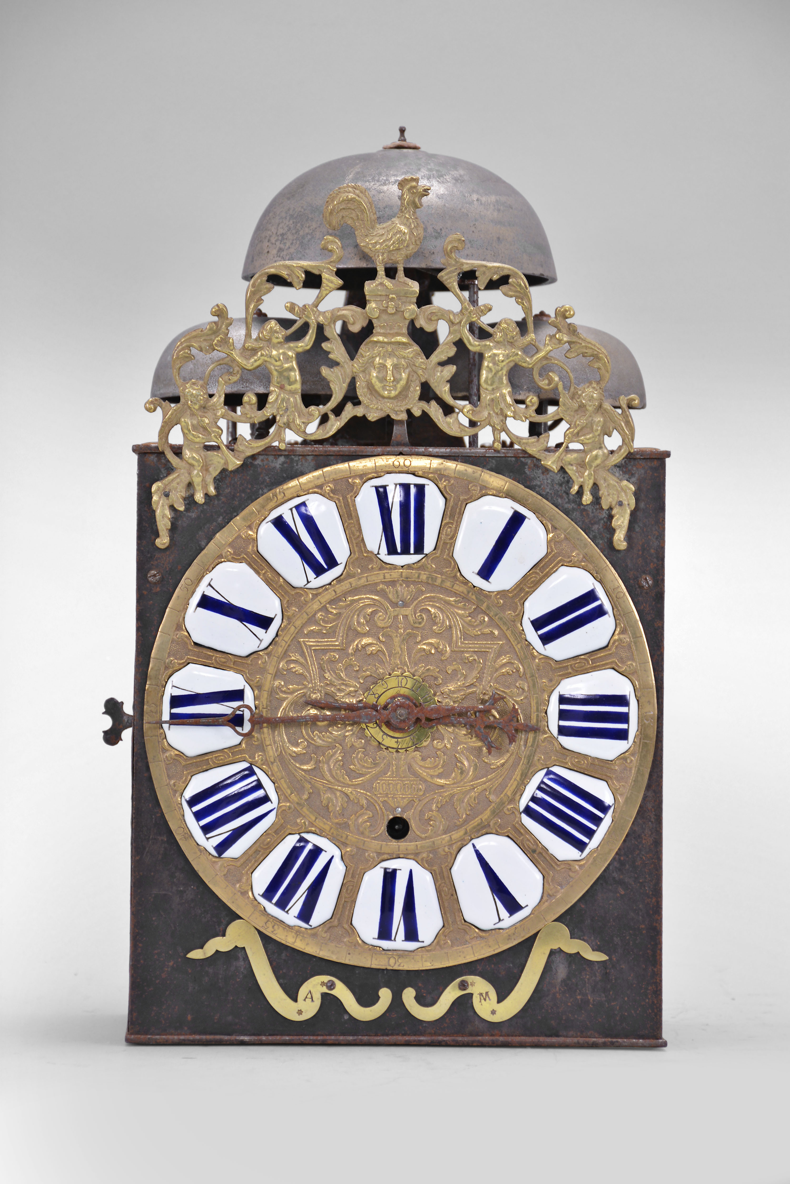 France, bronze-front Comtoise clock, 8 day, time and quarter strike with alarm, three bells, weight driven movement with verge escapement, thread suspension with rear-hanging pendulum in an iron post and plate frame, cast brass dial inset with 12 white enamel cartouches showing Roman numerals, Arabic numerals at 5 minute intervals on the outer rim, brass alarm ring with Arabic numerals, two iron hands, pivoted brass winding arbor shutters engraved with "A" and "M", all topped by cast fronton depicting trumpeters flanking a small forward facing Gallic rooster. — Antique Clocks Price Guide