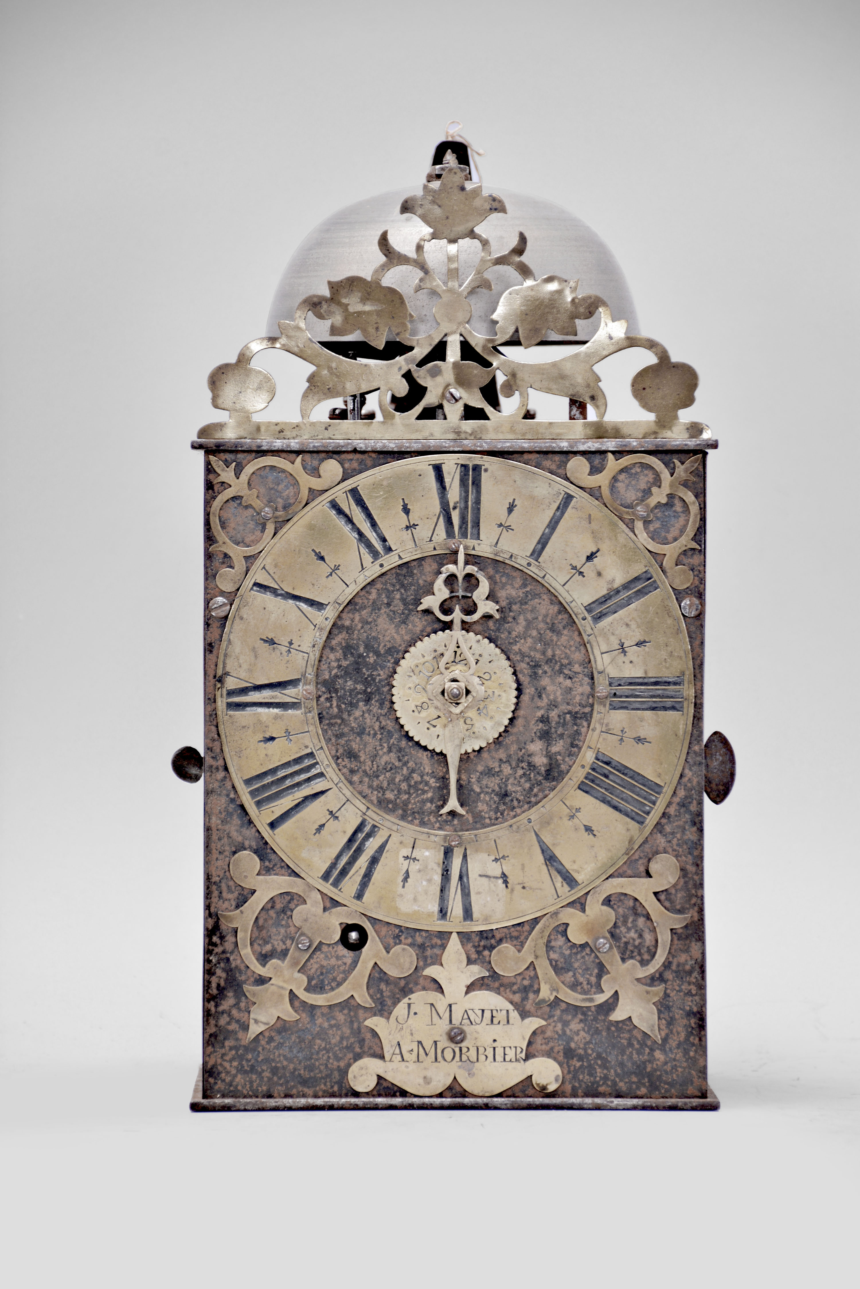 J. Mayet, Morbier, France, Comtoise clock, 8 day, time and alarm, weight driven movement with verge escapement, thread suspension with rear-hanging pendulum in a iron post and plate frame, brass chapter ring with engraved Roman numerals, brass alarm ring with engraved Arabic numerals, pierced brass spandrels and engraved plaque below the dial reading "J. Mayet a Morbier", single brass hand, all topped with a pierced brass fronton. — Antique Clocks Price Guide