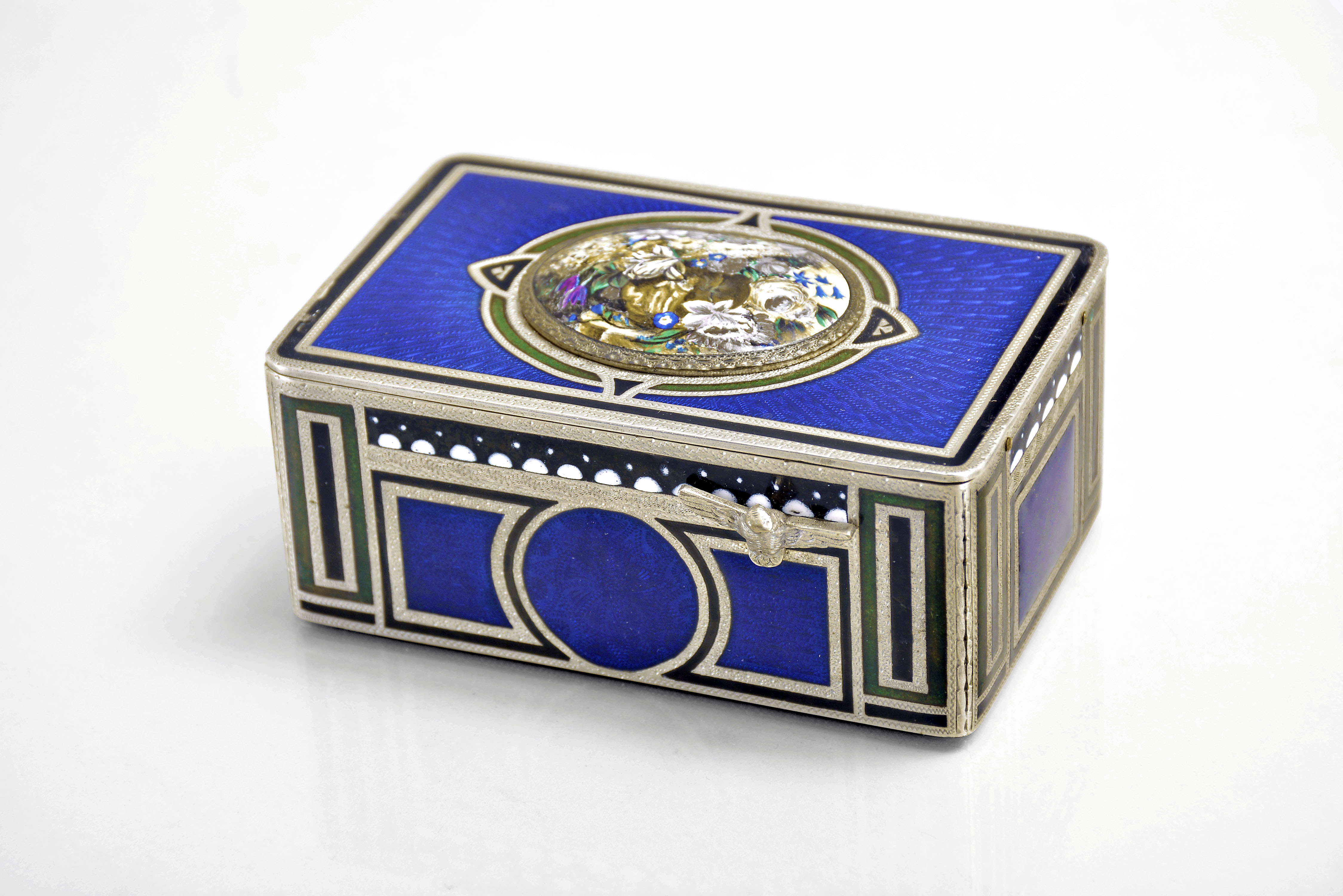 Karl Griesbaum, Germany, an enamelled silver singing bird box, the case with rectilinear panels decorated in blue, black, green and white enamels, the top in blue guilloche enamel with narrow black enamel border, the oval cover decorated with polychrome enamel flowers, and with green and black enamel border, opening to reveal a small, colorful bird, performing a short song and dance prior to disappearing beneath the cover, with key. — Antique Clocks Price Guide