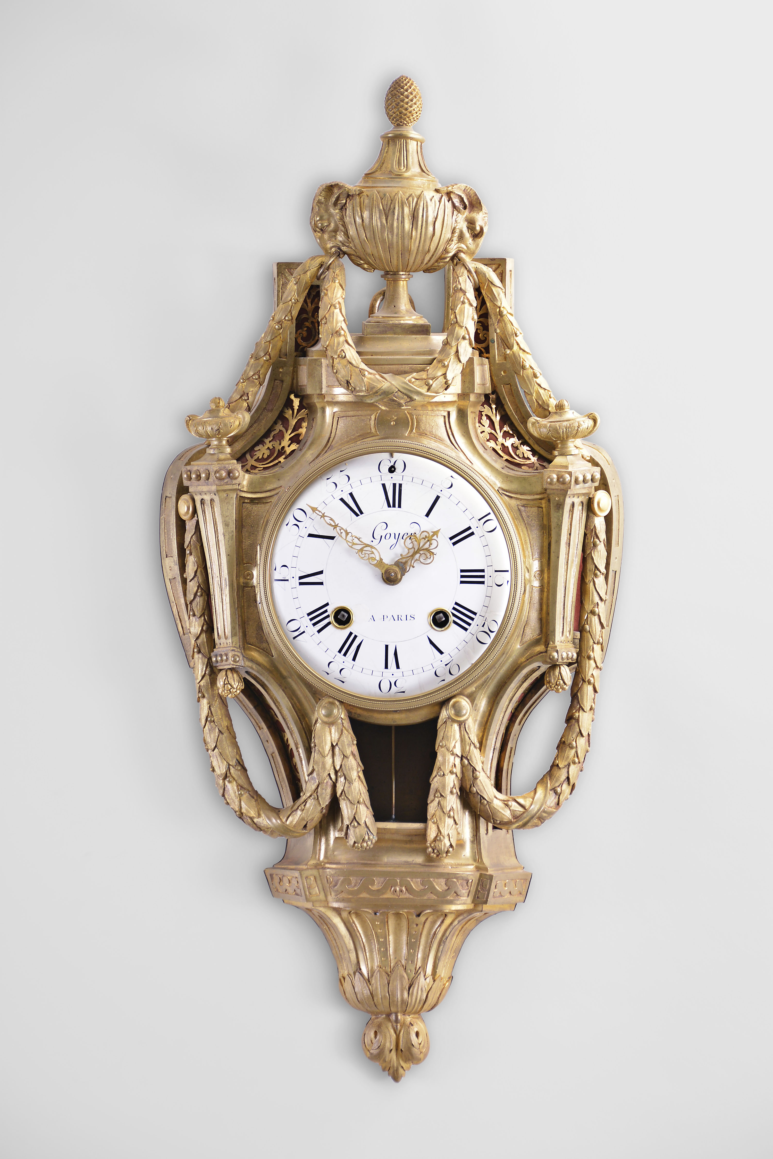 Goyer, a Paris, a neoclassical gilt bronze "Aux Guirlandes" cartel clock, the cast case surmounted by an urn with ram's heads at the sides, each with a ring in its mouth suspending a laurel festoon, the festoons continuing down the case sides and terminating beneath the bezel, case sides with textile backed apertures and foliate form brass frets, bottom with Vitruvian scroll panels above a fluted incurved bracket with laurel and acanthus ornament, roman numeral, convex white enamel dial with arabic five minute markers, signed "Goyer a Paris", well made pierced and engraved hands, large, flat bottomed round movement of at least two week duration, with recoil escapement, silk thread suspension and striking on a bell, the front plate with scribed circles for train layout, front and back plates also with holes for observing depth of engagemen between wheels and pinions — Antique Clocks Price Guide