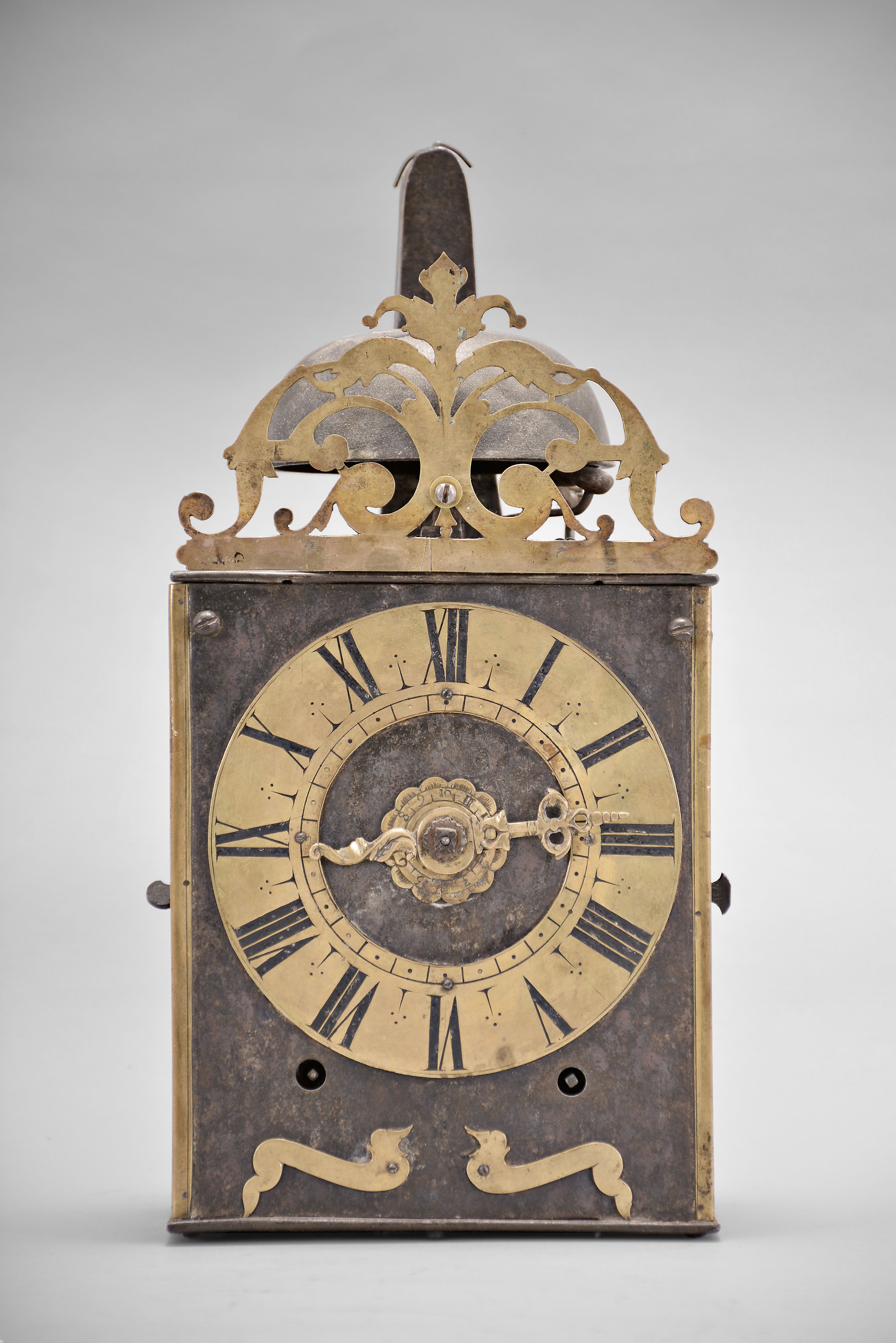 France, Comtoise clock, 8 day, time and strike with alarm, weight driven movement with verge escapement, thread suspension with rear-hanging pendulum in a iron post and plate frame, brass chapter ring with engraved Roman numerals, brass alarm ring with engraved Arabic numerals, pivoted winding arbor shutters, single pierced brass hand, the top with pierced brass fronton. — Antique Clocks Price Guide