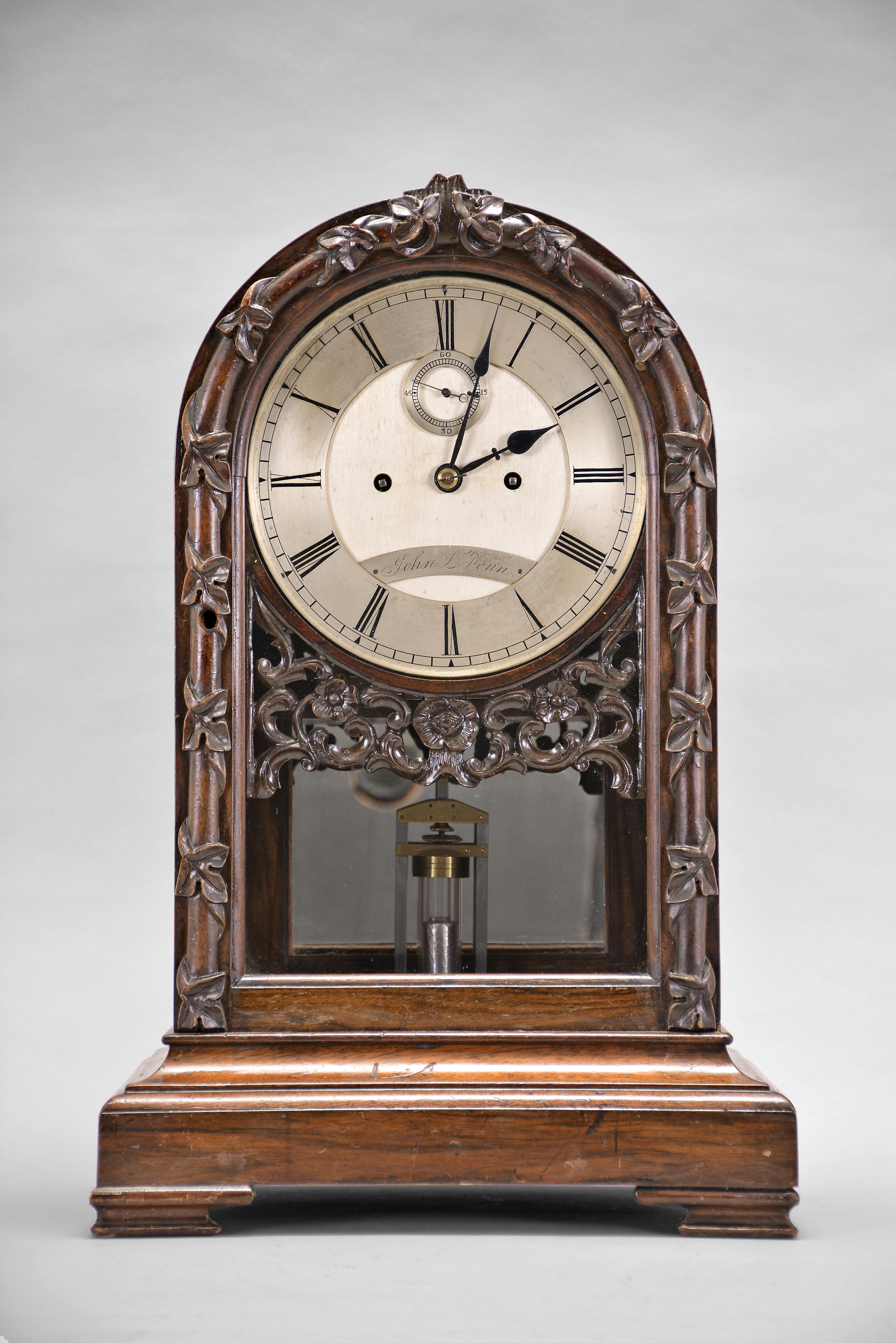 John L. Fenn, a Victorian mahogany table regulator, the arch top case with squat ogee bracket feet and molded base, the door with half round molding with carved leaves and vine, seemingly wrapped around the molding, Roman numeral silvered dial with seconds below 12:00, blued steel hands, and with pierced floral and C scroll carving below, large 8 day fusee movement striking the hours on a coil gong, faux mercury pendulum with heavy brass cylinder fixed horizontally below the jar — Antique Clocks Price Guide